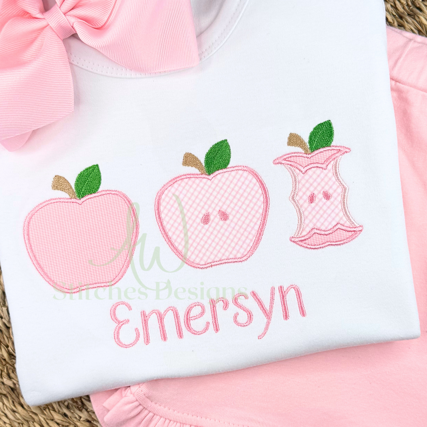 White shirt with pink embroidered apples and text, placed on a pink surface.
