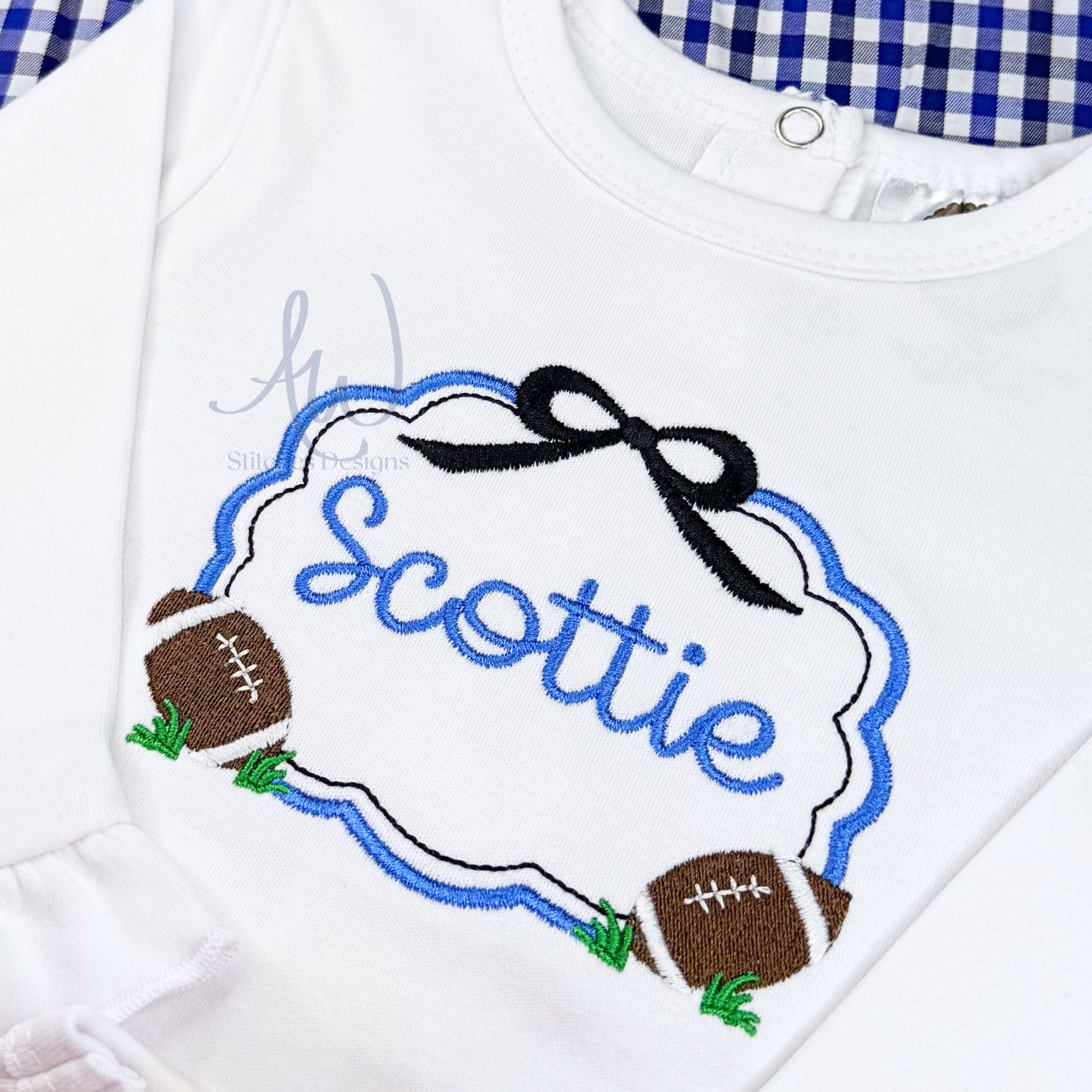 A close-up of a white long-sleeve baby or toddler top with a custom embroidered design. The design features a blue and white cloud outline with the name 'Scottie' in blue script, a black bow at the top, and two brown footballs with green grass accents on either side.