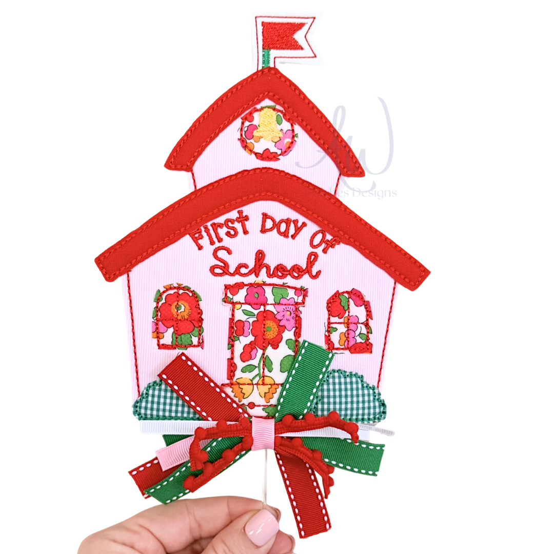 A handmade embroidered schoolhouse ornament with the words 'First Day of School' in red script. The schoolhouse features red trim, a pink body, floral window patterns, a small flag on top, and a decorative red and green ribbon bow at the bottom.
