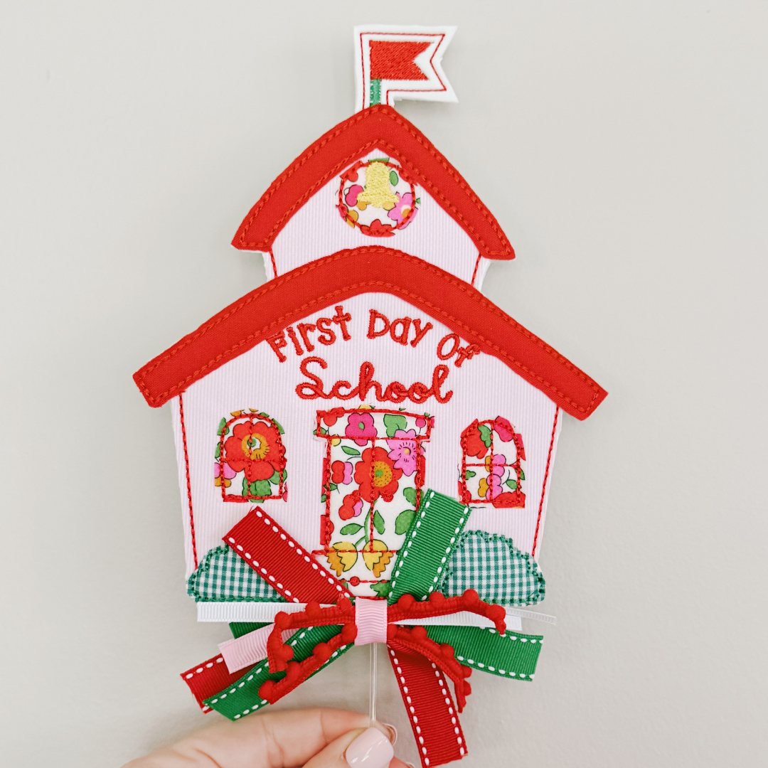 A handmade fabric schoolhouse decoration with 'First Day of School' text, floral details, and ribbon bows, held by a hand against a plain light background.