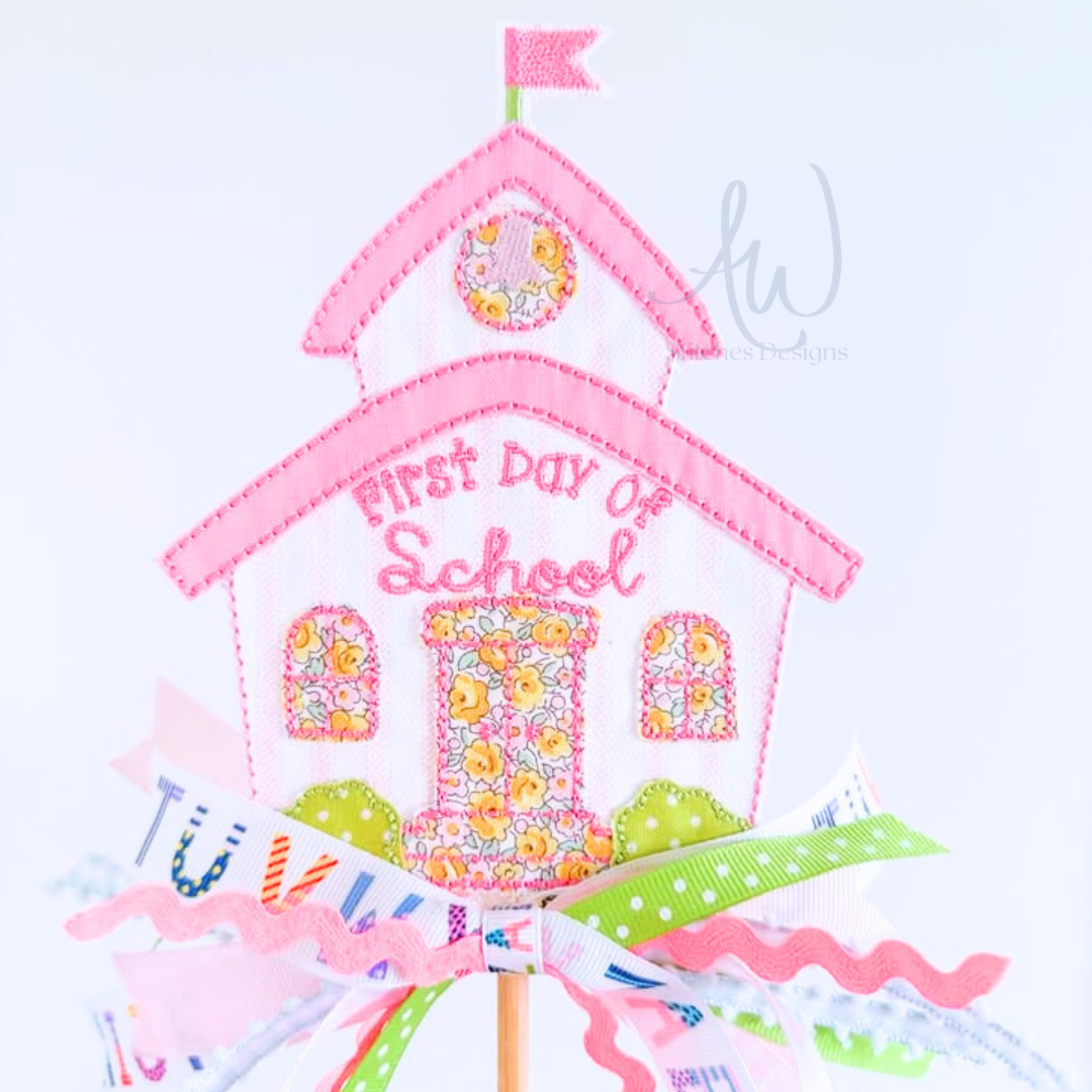 A decorative schoolhouse-shaped cake topper or centerpiece with pink embroidered text reading 'First Day of School,' floral patterned details, ribbon bows, and a small flag on a stick against a light background.