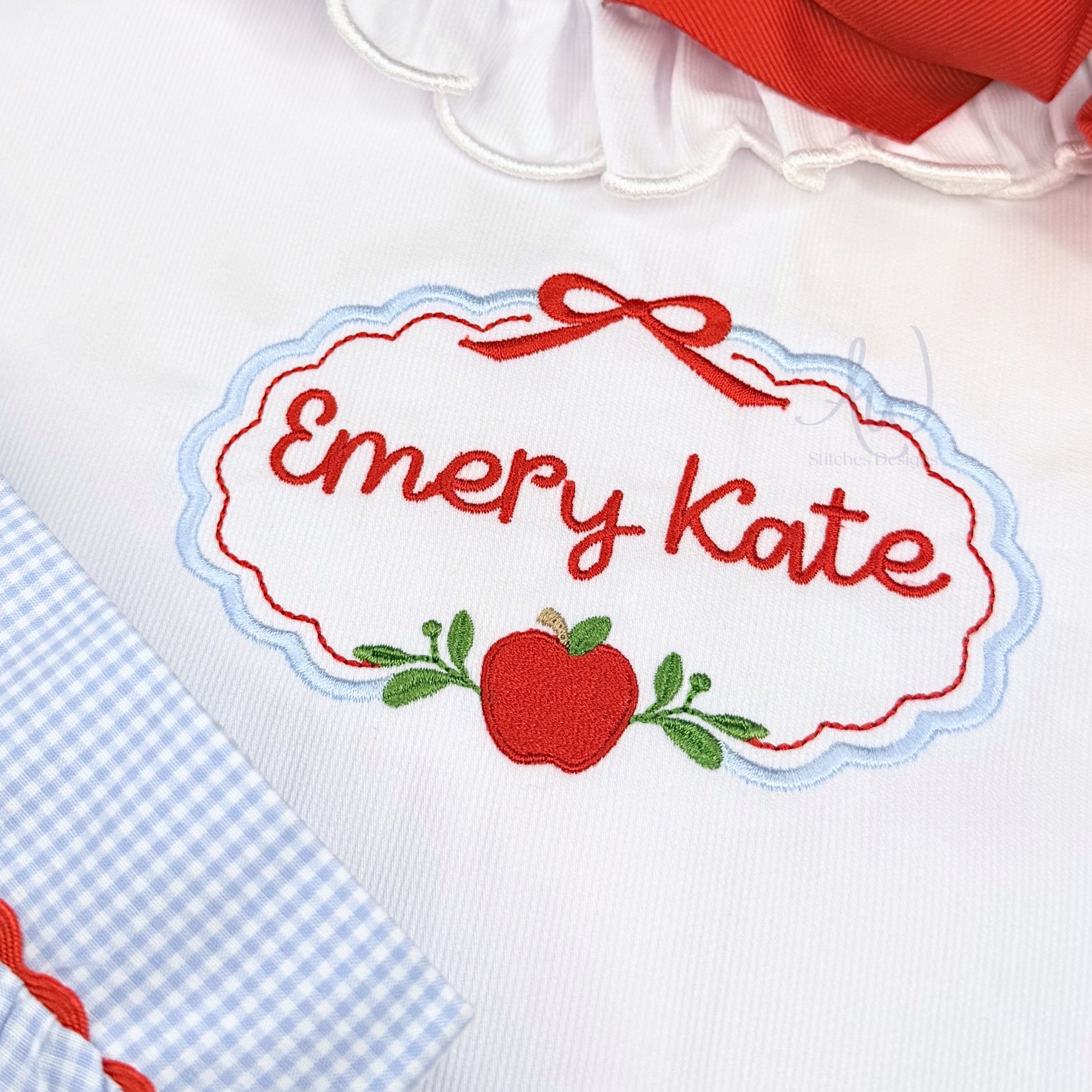 Close-up of a white embroidered patch with the personalized name 'Emery Kate' in red script, surrounded by a red ribbon and red apple with green leaves. Part of a blue-and-white gingham fabric and a red ribbon are visible at the edges.