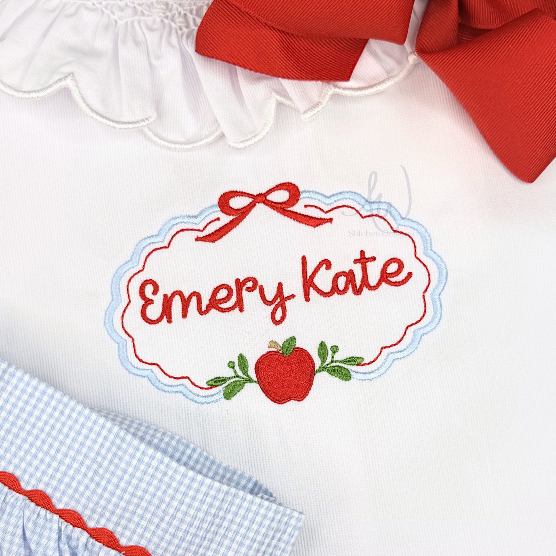 Close-up of a white baby dress with red and blue embroidery reading 'Emery Kate' around an apple motif, plus a red bow at the neckline and a blue-and-white gingham skirt edge.