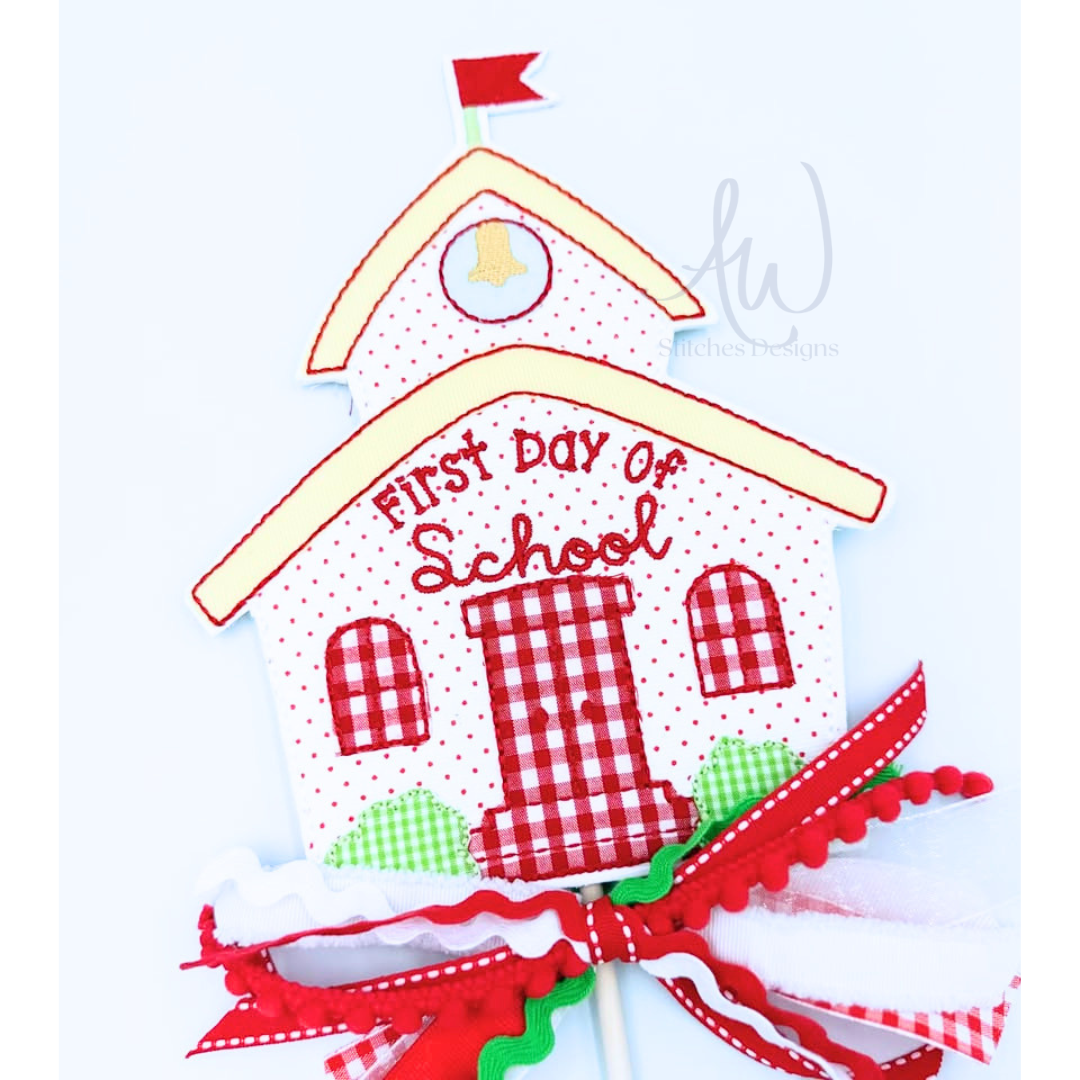 A handmade schoolhouse-shaped first day of school cake topper or decoration on a white stick, featuring red and white gingham patterns, red script text reading 'First Day of School,' a small red flag, and a large red, white, green, and white ribbon bow at the bottom.