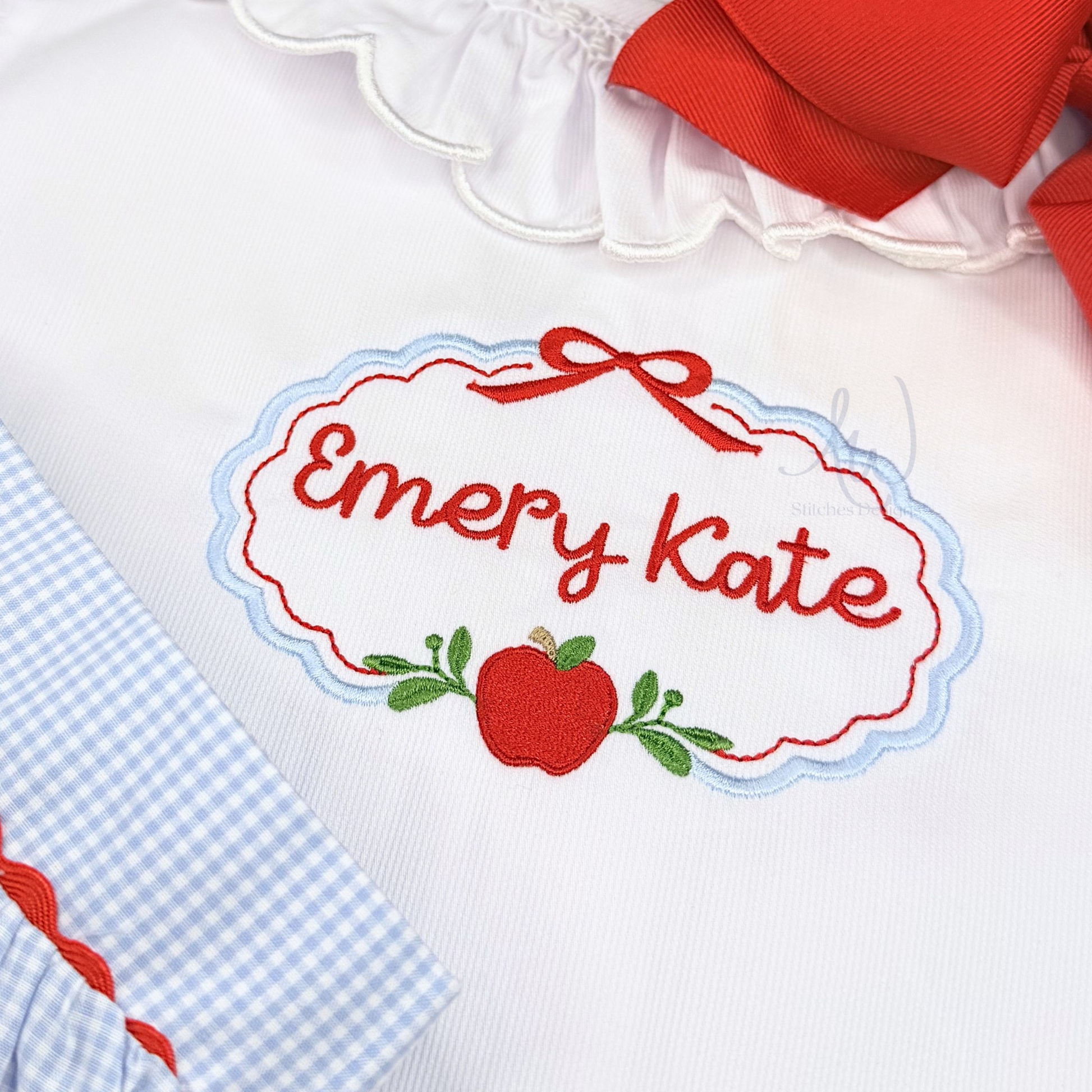 Close-up of a white embroidered nameplate applique reading 'Emery Kate' in red script, outlined with a red and light blue border and topped with a red ribbon motif. A red apple with green leaves is stitched below.