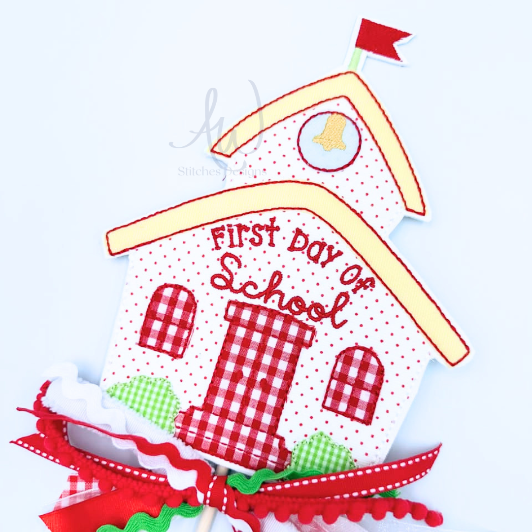 A decorative schoolhouse-shaped applique or patch with the words “FIRST DAY OF School” stitched on it. The design features red and white gingham patterns, red polka dots, a red bell, a small red flag, green gingham bushes, and red-and-white checkered windows.