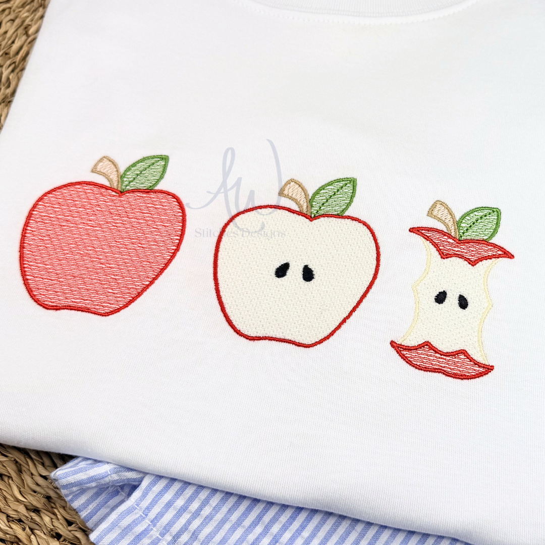 A close-up of a white embroidered baby shirt or blanket featuring three cute apple designs: a whole red apple, a sliced apple showing seeds, and a bitten apple with a red bite taken out. The shirt is laid flat on a woven mat with a blue and white striped fabric underneath.
