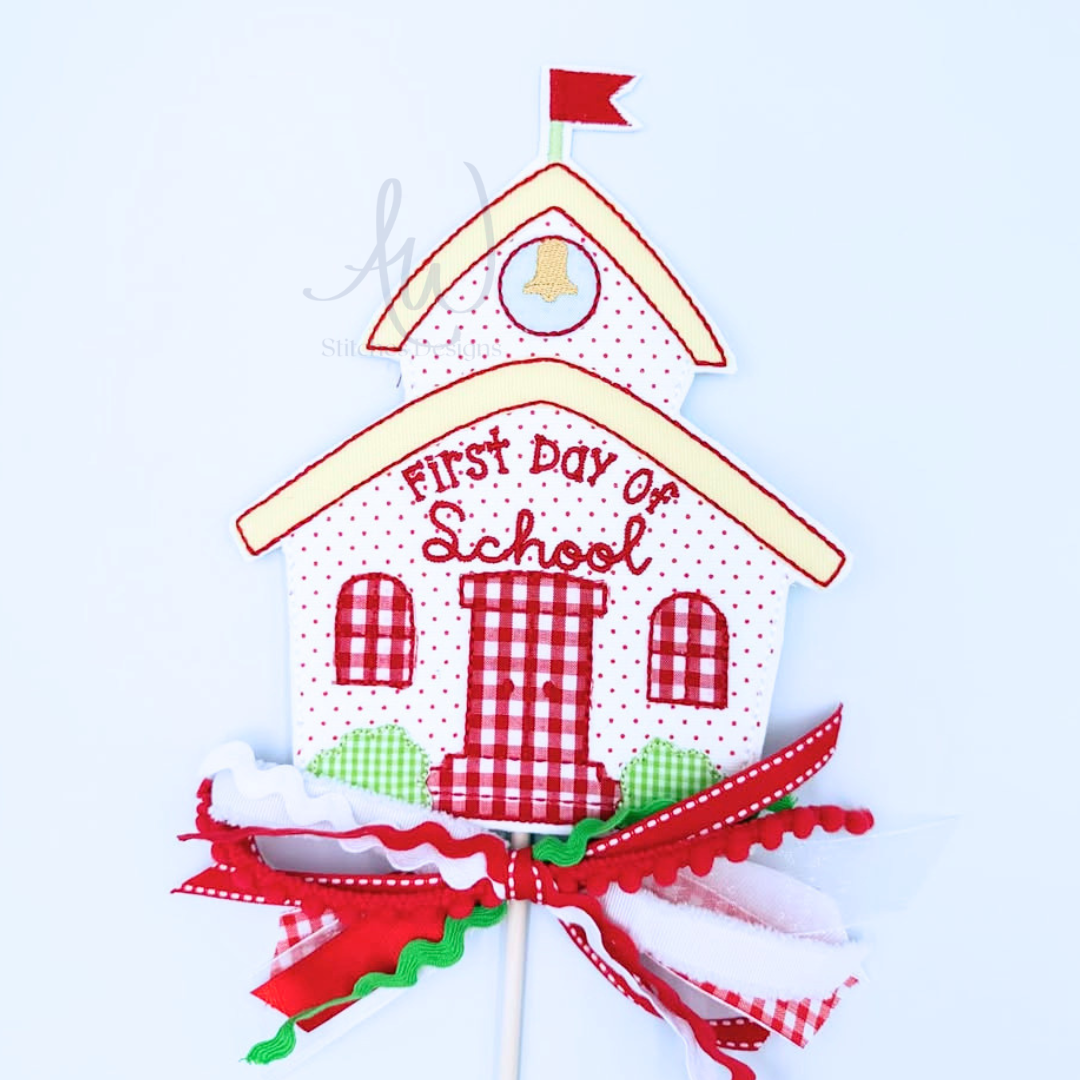 A decorative schoolhouse-shaped cake topper or party decoration with the words “First Day of School” on the front, featuring red and white gingham patterns, a red ribbon bow at the bottom, and a small red flag on top, shown against a plain light background.