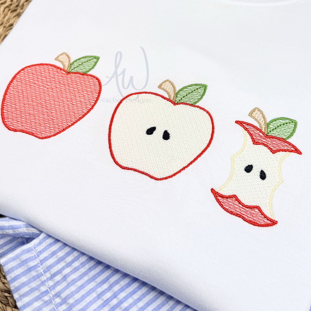A close-up of a white garment featuring embroidered apple motifs: a whole red apple, a sliced apple with black seeds, and an apple core with red lips. A blue-and-white striped fabric and woven mat are partially visible underneath.