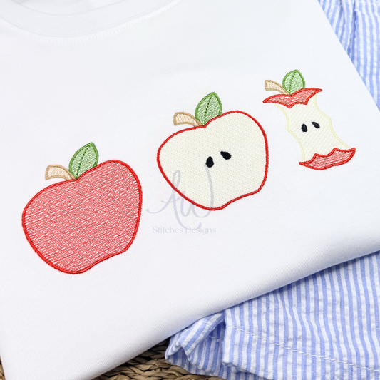 A close-up of a white garment featuring embroidered apple motifs: a whole red apple with green leaves, a sliced apple showing cream flesh with black seeds, and an apple core. Folded blue-and-white striped fabric and a woven surface are visible underneath.