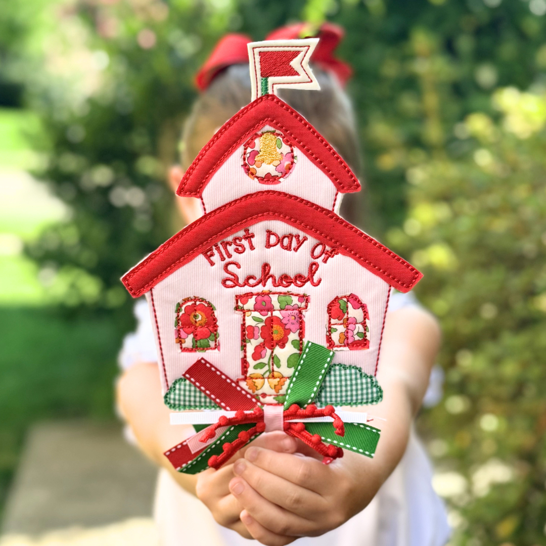A child holds up a decorative house-shaped applique or patch that reads "First Day of School" with red trim, floral details, and ribbon bows, against a blurred outdoor garden background.