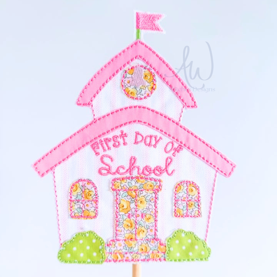Embroidered schoolhouse applique patch with 'First Day of School' text, pink trim, floral patterned windows and door, green bushes, and a small pink flag on a wooden stick, shown on a light gray background.