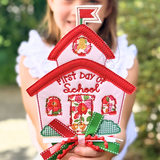 A person holding a decorative house-shaped patch that reads 'First Day of School' with red trim, floral details, and a ribbon bow, against a blurred outdoor background.