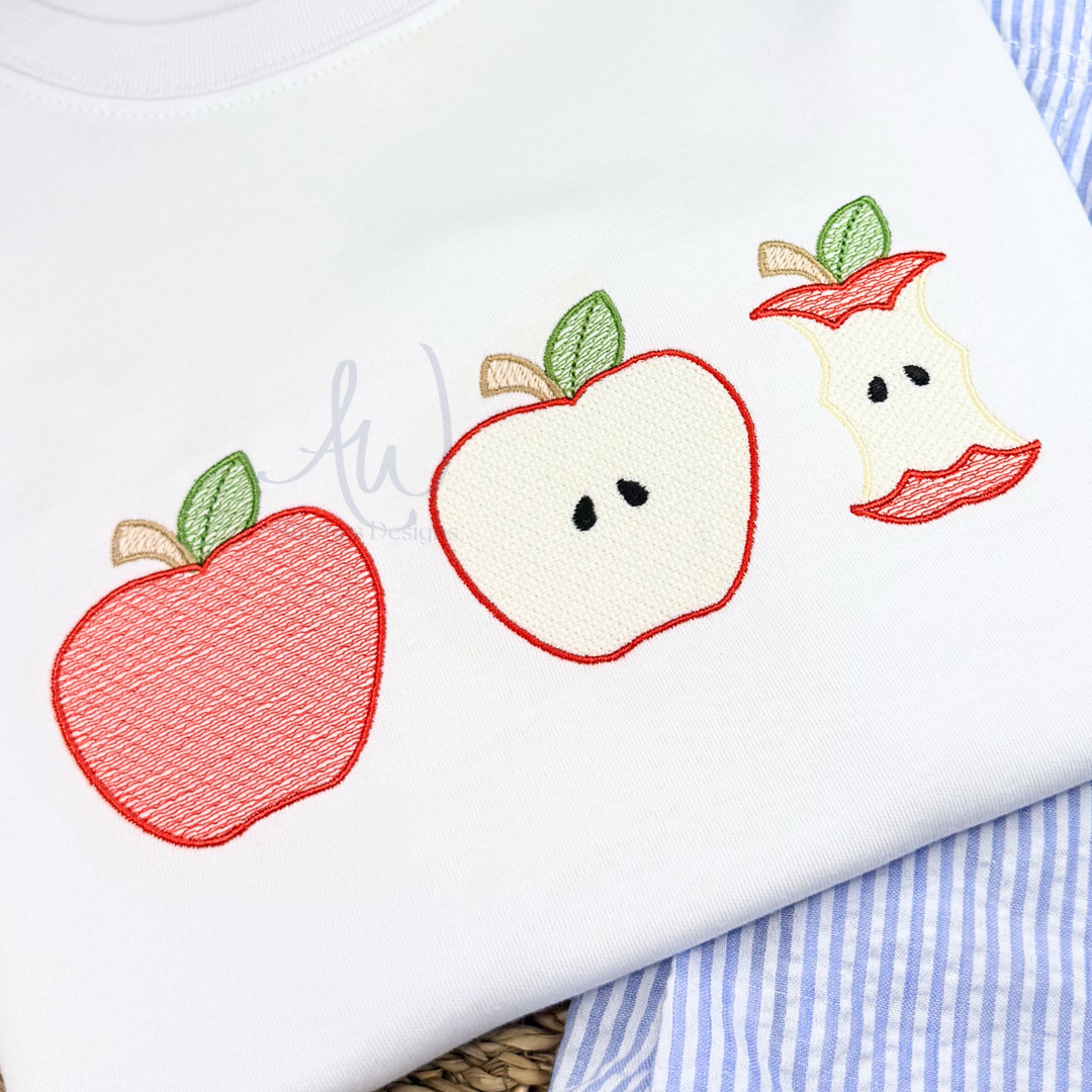 A close-up of a white garment featuring embroidered apple motifs in red, cream, green, and tan. A blue-and-white striped fabric and a woven surface are visible beneath the garment.