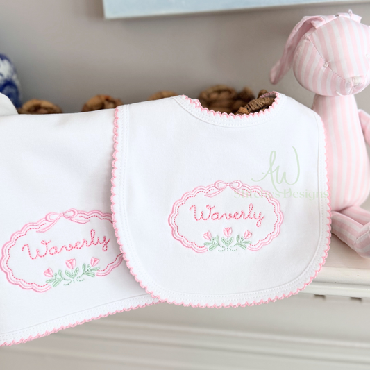 White baby bib and burp cloth featuring the Rosebud Floral Embroidery Frame Design with pink floral border, bow detail, and tulip accents by AW Stitches Designs