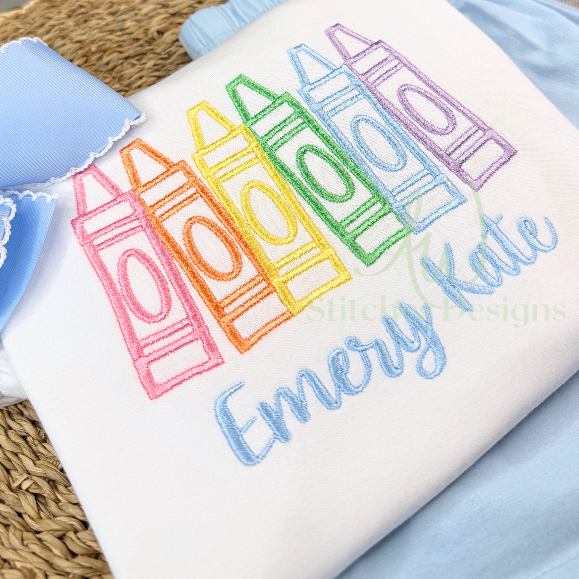 Embroidered design of colorful crayons with the name 'Emery Kate' on a white fabric background.