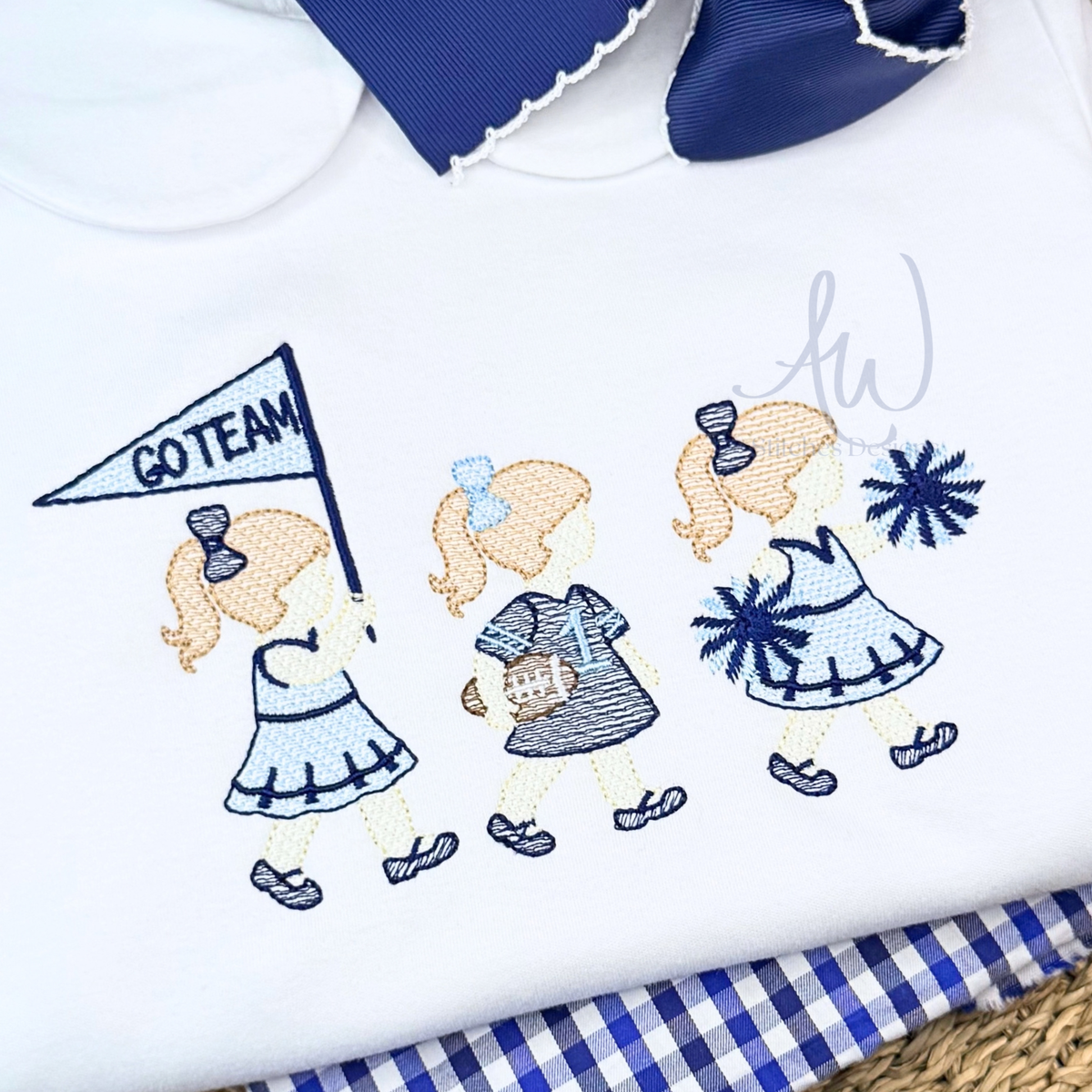 A close-up of a white embroidered t-shirt featuring a cheerleader girl graphic holding a 'GO TEAM' pennant, styled with a blue ribbon and a blue-and-white gingham check fabric underneath.
