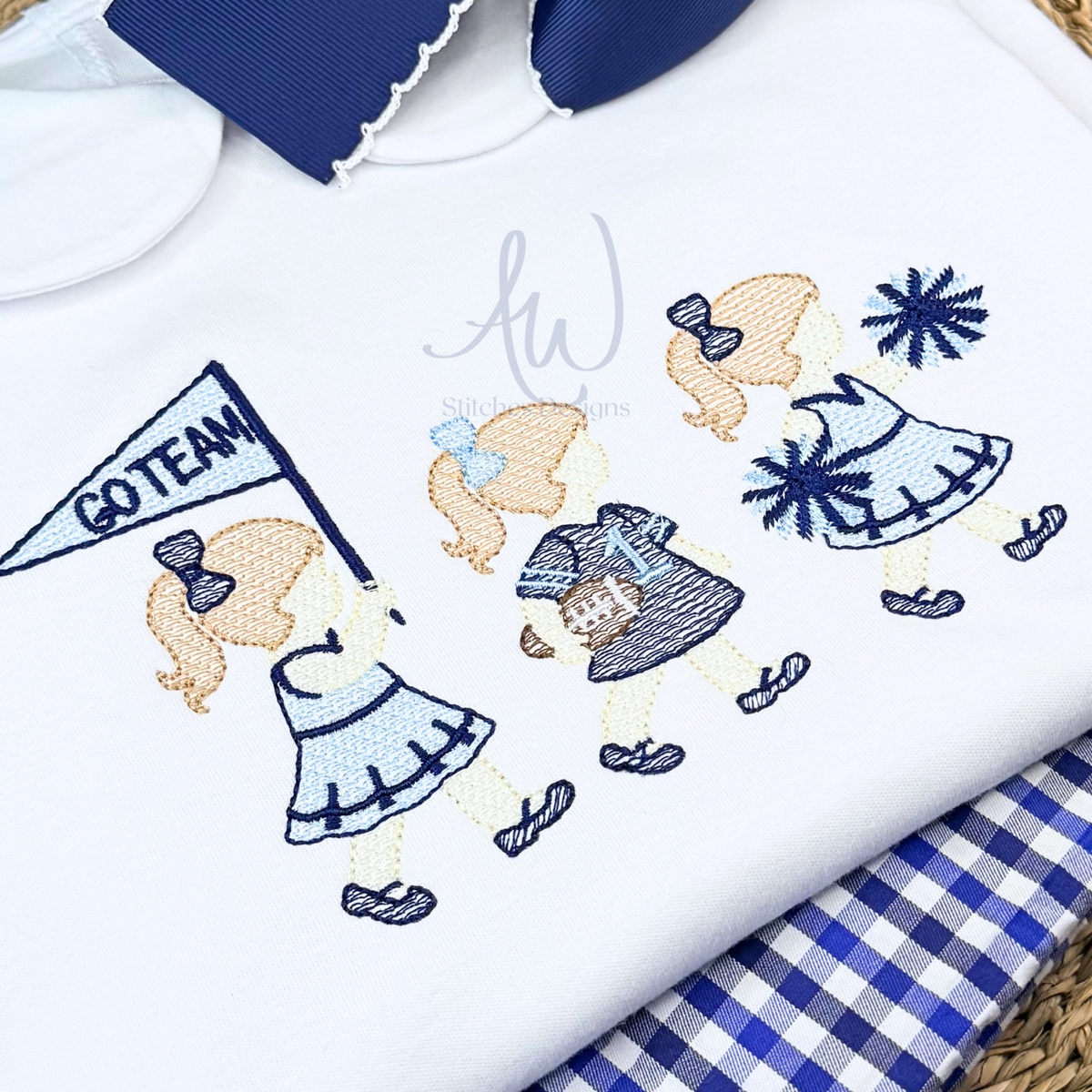 A close-up of a white embroidered dress featuring three cheerleader girls in blue and navy outfits, one holding a 'GO TEAM!' flag, one holding a football and one holding pom poms.