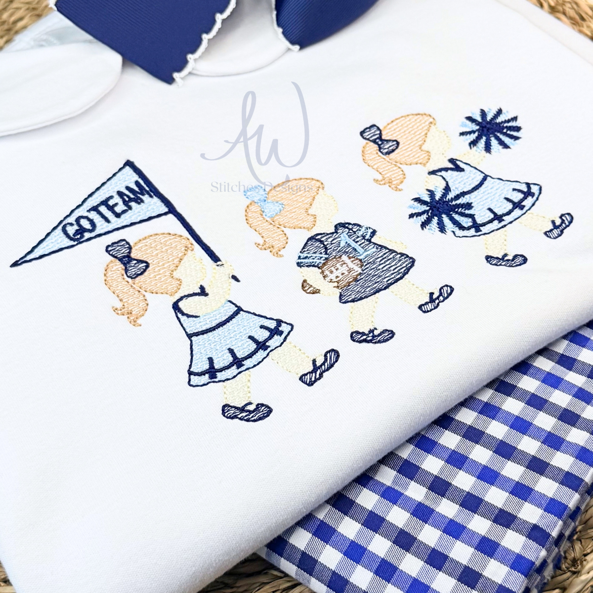 A folded white dress embroidered cheerleader girls in blue and navy, including a flag that says GO TEAM, layered over a blue-and-white gingham fabric. 