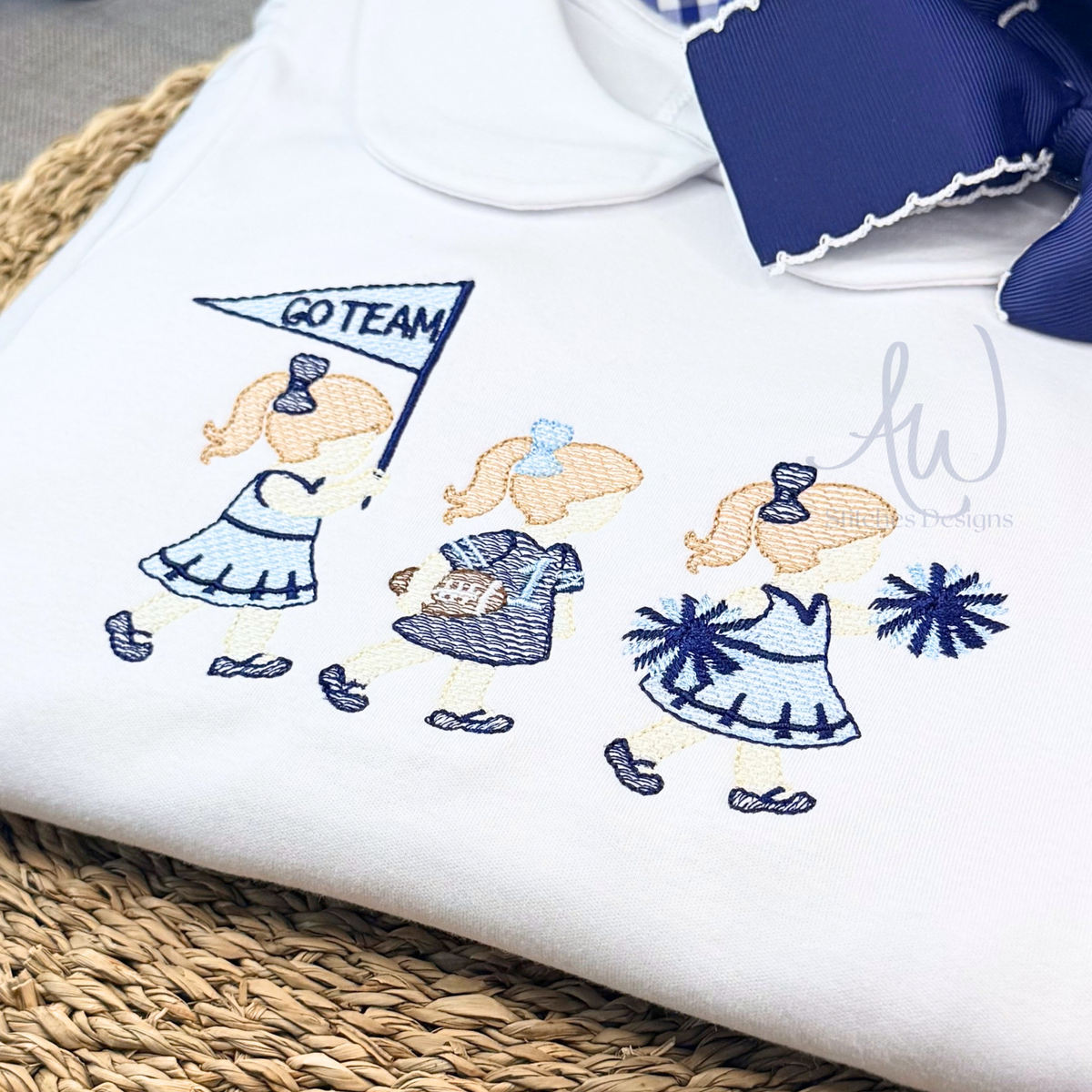 A folded white dress featuring embroidered cheerleader girls in blue and navy dresses, one holding a 'GO TEAM' flag, with a navy blue ruffled trim and bow at the top. 