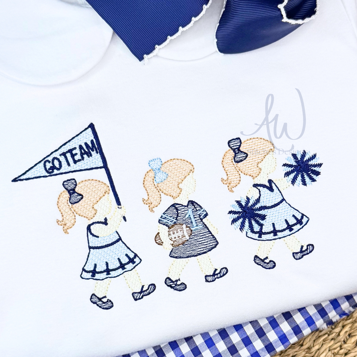 Close-up of a white embroidered dress featuring three girls dressed in blue and navy outfits, one holding a 'GO TEAM' pennant, with a blue bow and blue gingham fabric visible around it.