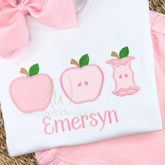 White shirt with pink embroidered apples and text, placed on a pink surface.