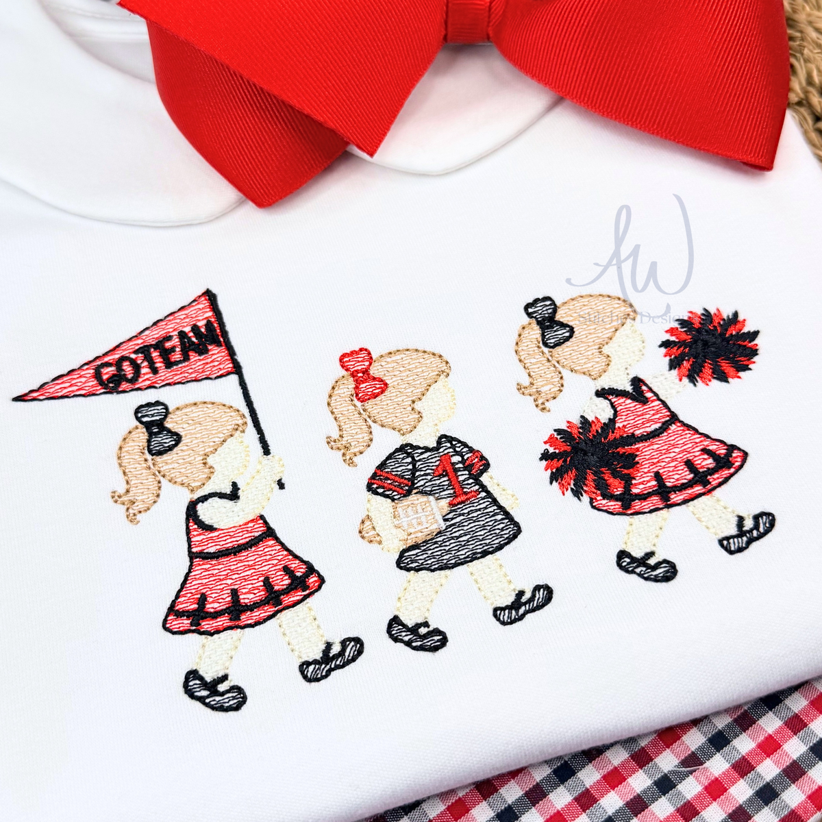 A close-up of a white embroidered T-shirt featuring three cartoon girls in red and black outfits, with a large red bow at the collar. The shirt has a visible 'AW' monogram embroidery.