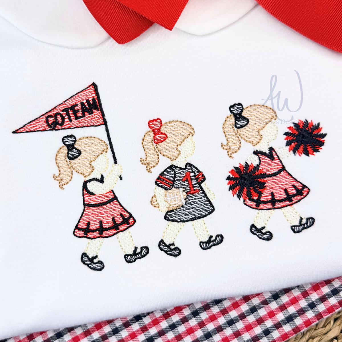 A close-up of a white embroidered baby garment, likely a shirt or bodysuit, featuring three cartoon cheerleader girls walking. One girl holds a pink 'GOTENN' flag, and two others wear red and black pom-poms.