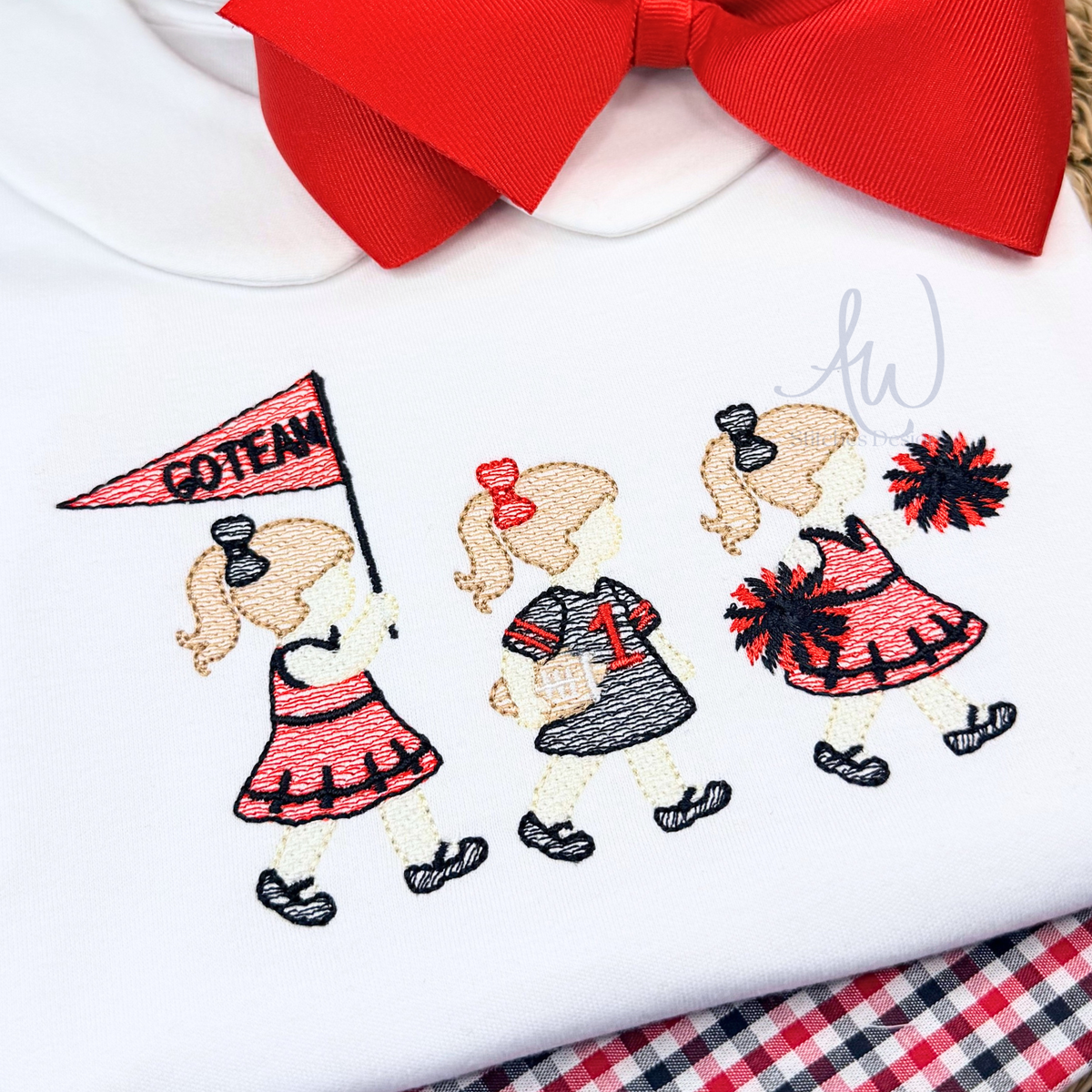 A close-up of a white baby garment featuring embroidered girls in cheerleader outfits, with a large red bow at the collar and a red-and-white plaid fabric visible underneath.