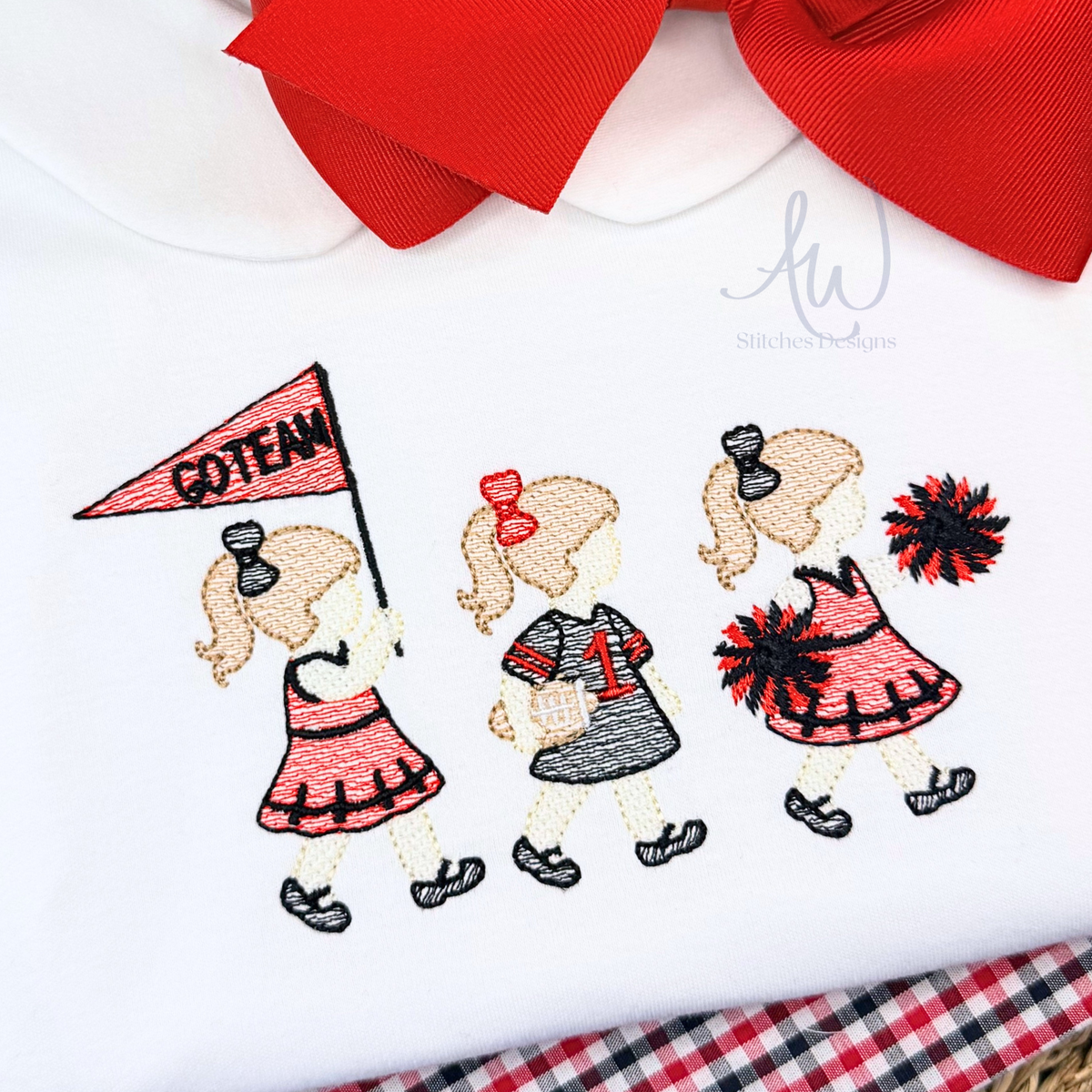 A close-up of a white embroidered garment featuring three cartoon cheerleaders in red, pink, and gray outfits, with one holding a pink 'GO TEAM' flag. A large red bow is partially visible at the top, and a red-and-black plaid fabric edge appears at the bottom right.