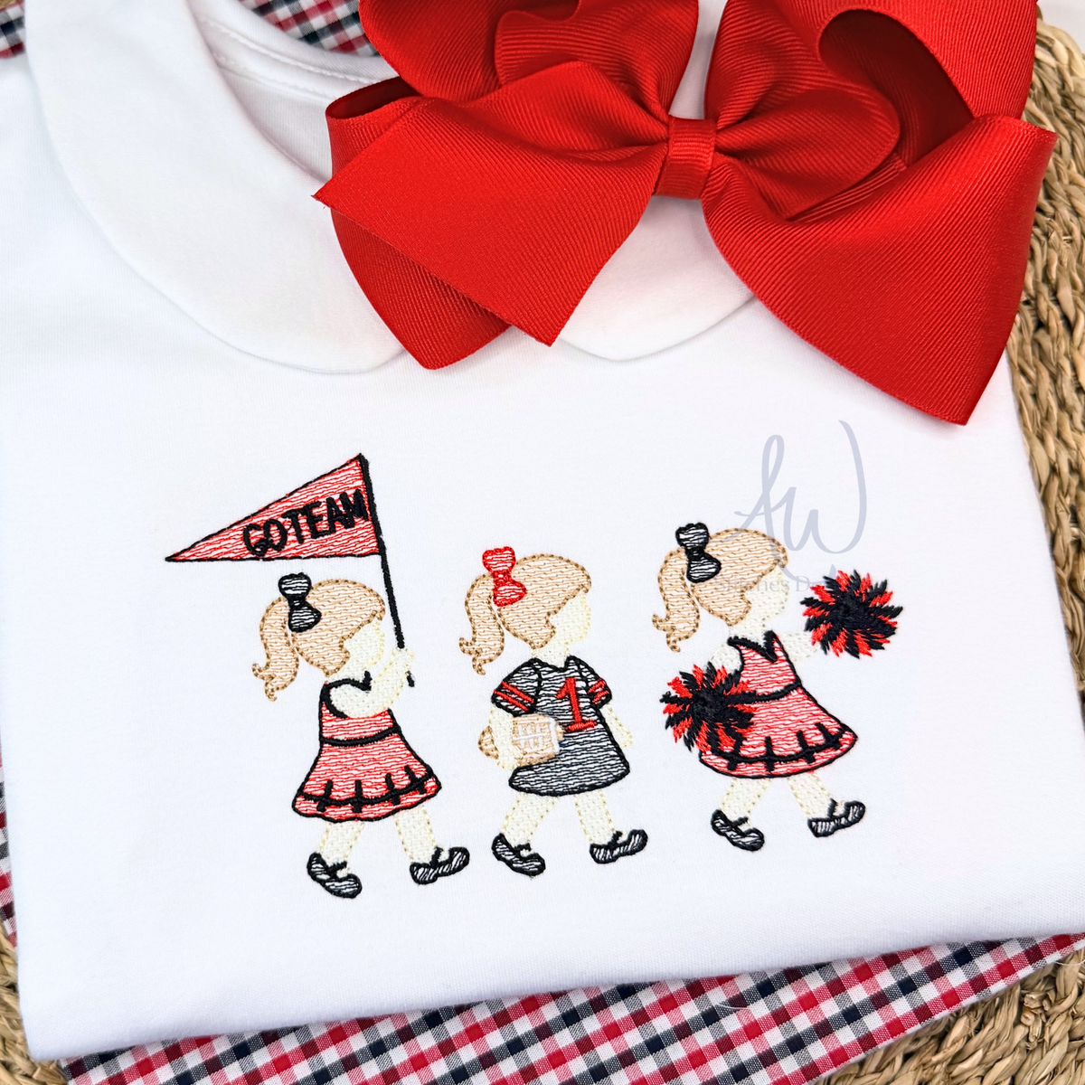 A white baby or toddler long-sleeve shirt laid flat on a red-and-white plaid blanket and woven mat, featuring embroidered cheerleaders and a large red satin bow at the neckline.